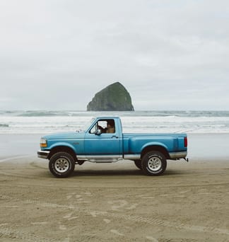 A blue pickup truck parked on the beach at Pacific City, with Haystack Rock behind it.